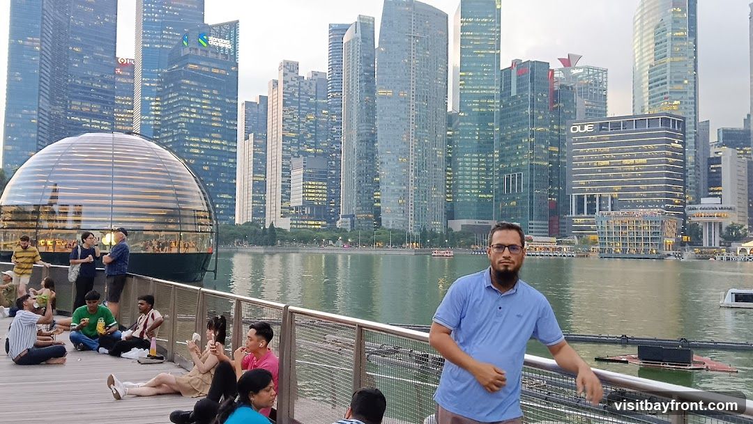 A group of people are sitting on a wooden deck by a body of water. In the background, a modern cityscape with numerous skyscrapers is visible. A distinctive glass dome structure is also prominent in the scene. A man in a blue polo shirt is standing in the foreground, looking towards the camera.