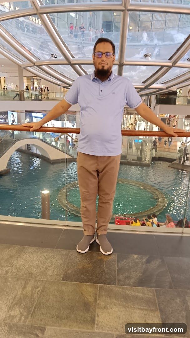A man stands with his arms outstretched, posing for a photo in what appears to be a large indoor shopping mall. Behind him, a multi-level atrium features a water feature with a bridge and people in boats. The architecture includes a modern, curved glass ceiling and metal supports. Various shops and escalators are visible in the background on different levels.