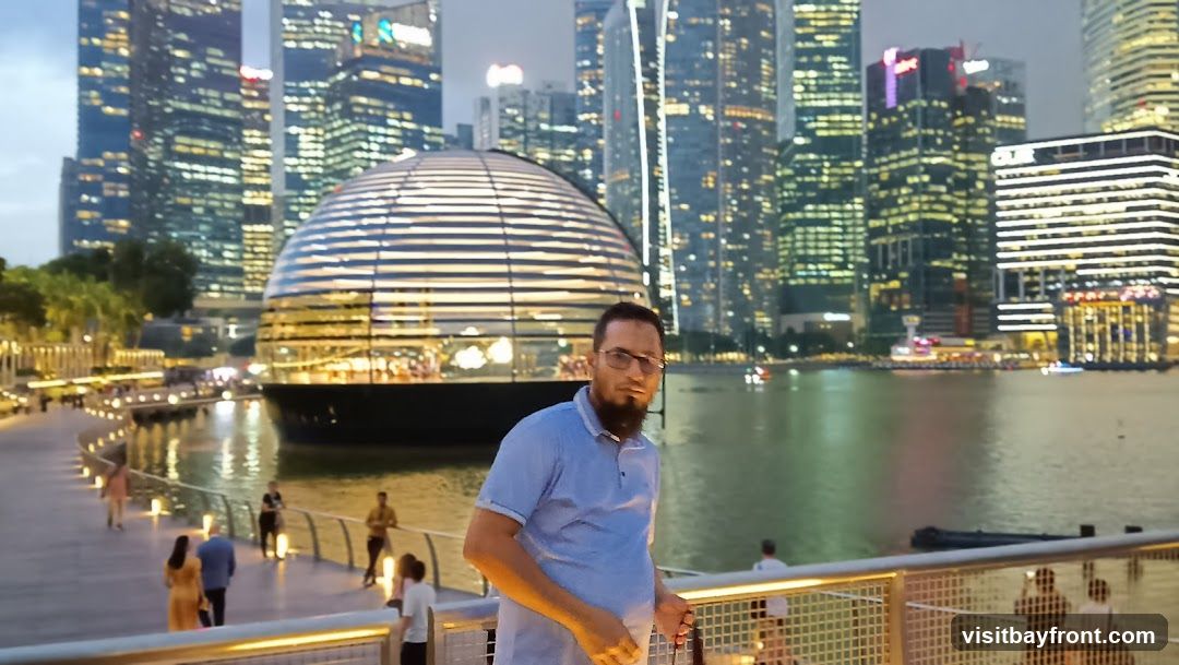 A man wearing a light blue polo shirt and glasses stands on a walkway, with a distinctive glass dome building behind him. The background features a cityscape with numerous illuminated skyscrapers at dusk. People are visible on the walkway and in the distance, and boats are on the water.