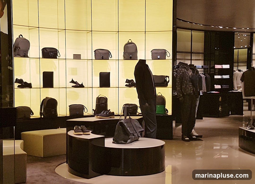 A well-lit interior of a luxury store featuring a display of handbags, shoes, and clothing on mannequins. The shelves are illuminated with a warm yellow light, showcasing various bags and footwear. In the foreground, a large black handbag is displayed on a circular pedestal. Further back, mannequins are dressed in stylish attire, and racks of folded shirts are visible.