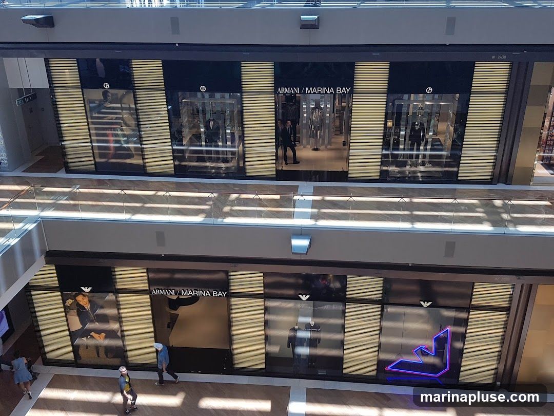 An aerial view of the Armani store at Marina Bay. The storefront features large display windows showcasing mannequins in suits and casual wear. The Armani logo and "Armani / Marina Bay" text are visible on the facade. The store has a modern design with golden slatted panels.