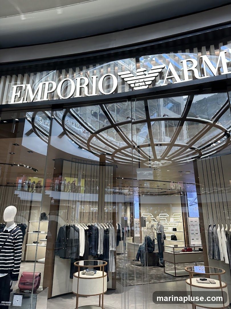 The exterior of an Emporio Armani store, viewed through a large glass window. The prominent white text 'EMPORIO ARMANI' with the iconic eagle logo is displayed above the entrance. Inside, mannequins are dressed in clothing, and racks of apparel are visible, along with display shelves and furniture. The store appears to be located in a modern shopping mall with a circular ceiling structure overhead.