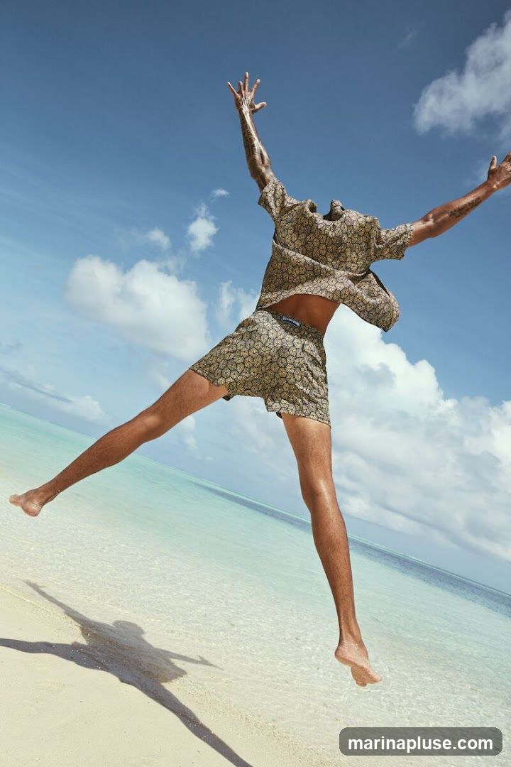 A man is captured mid-air, jumping with his arms outstretched against a bright blue sky with scattered clouds. He is wearing a patterned short-sleeved shirt and matching shorts. The scene is set on a sandy beach with clear turquoise water in the background.