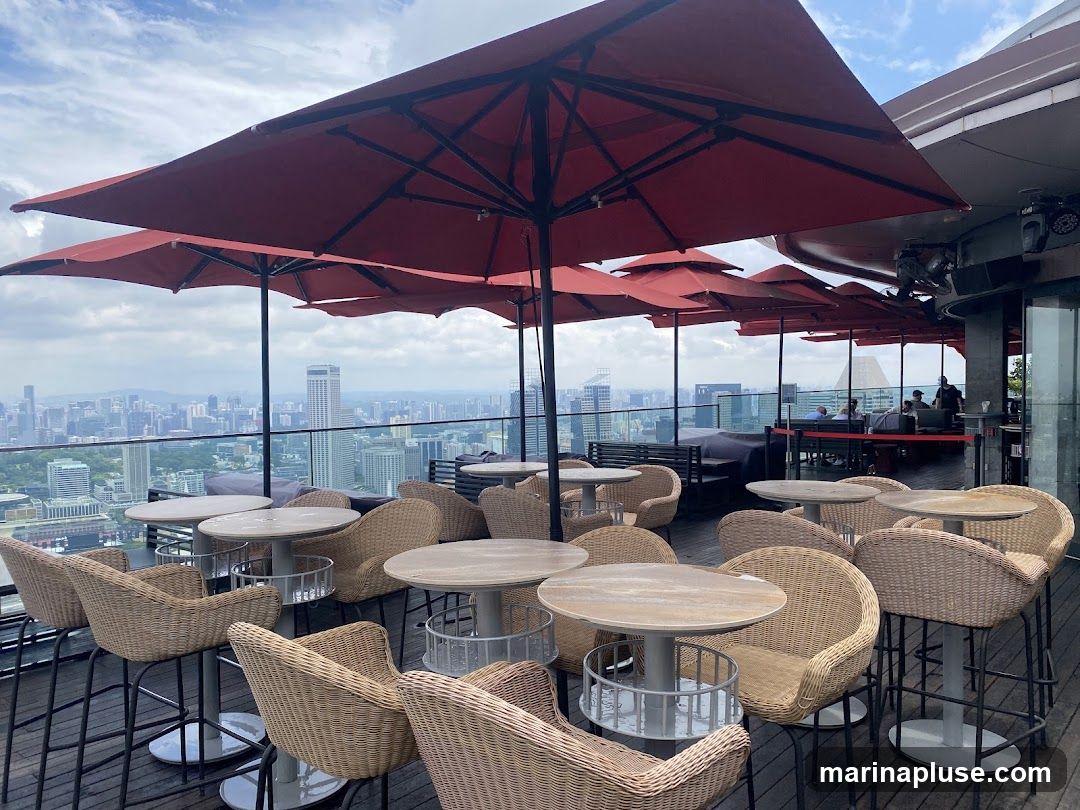 An outdoor seating area on a rooftop, featuring numerous round tables and wicker chairs. Red umbrellas provide shade over the tables. In the background, a sprawling cityscape is visible under a cloudy sky. Several people are seated at tables in the distance, and some bar equipment is visible on the right.