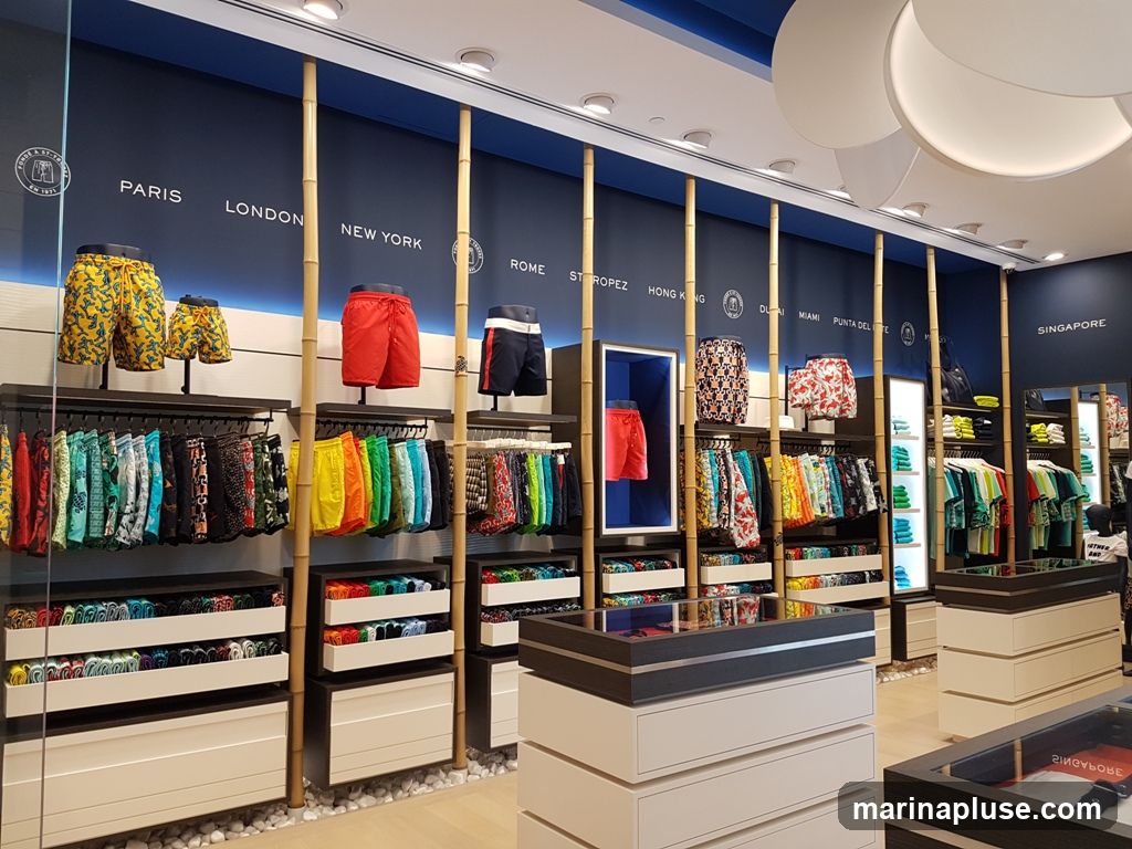 A well-lit interior of a swimwear store. The walls are painted navy blue with white text listing various cities like Paris, London, New York, Rome, St. Tropez, Hong Kong, Dubai, Miami, Punta del Este, and Singapore. Bamboo poles are used as decorative elements and supports for shelving. The store displays a wide variety of colorful swim trunks and shorts on racks and shelves. Some swim trunks are also displayed on mannequins or in illuminated display boxes. Below the hanging racks, there are drawers and shelves filled with folded swimwear. In the foreground, there are display counters with glass tops, showcasing more merchandise.