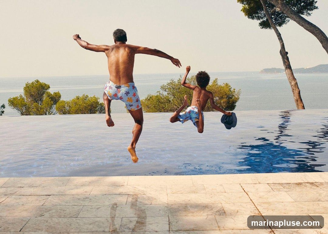 A man and a young boy are captured mid-air as they jump into an infinity pool. The man, wearing patterned swim trunks, is on the left, with his arms outstretched. The boy, also in patterned swim trunks, is on the right, holding a dark hat. The pool overlooks a scenic ocean view with lush green trees and a clear sky. The poolside is paved with large, light-colored tiles.
