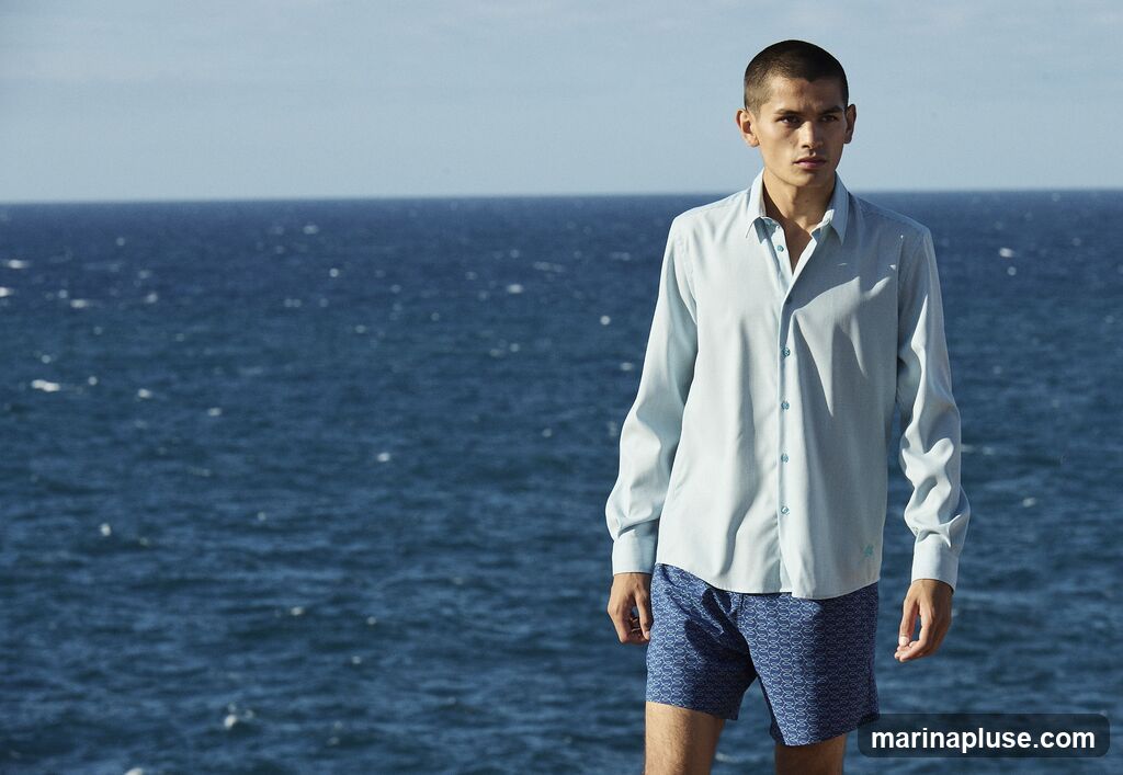 A young man stands against a backdrop of the blue ocean and sky. He is wearing a light blue, long-sleeved collared shirt and blue patterned shorts. The shirt is buttoned up, and the shorts have a repeating wave-like pattern.