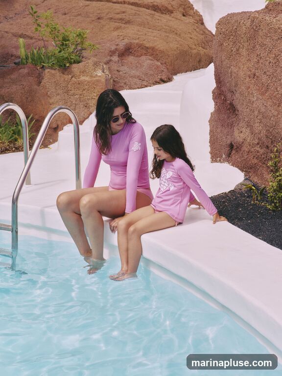 A woman and a young girl are sitting on the edge of a swimming pool, with their feet dangling in the water. They are both wearing pink long-sleeved rash guards. The woman has sunglasses on and her hair is dark and wavy. The girl's hair is also dark and she is looking down. The pool is filled with clear blue water. The background features rocky, reddish-brown terrain and white structures.