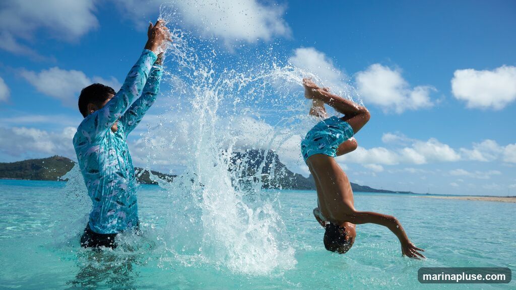 A person in a long-sleeved rash guard with a fish pattern splashes water upwards, while another person in patterned swim trunks performs a backflip in the turquoise ocean. The sky is blue with scattered white clouds, and a mountainous island is visible in the background.