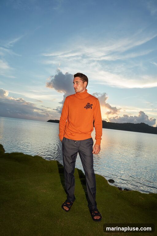 A man stands on a grassy hill overlooking a body of water at sunset. He is wearing an orange turtleneck sweater with a dark graphic design on the chest, dark pants, and dark sandals. The sky is filled with clouds and the sun is setting, casting a warm glow over the scene.