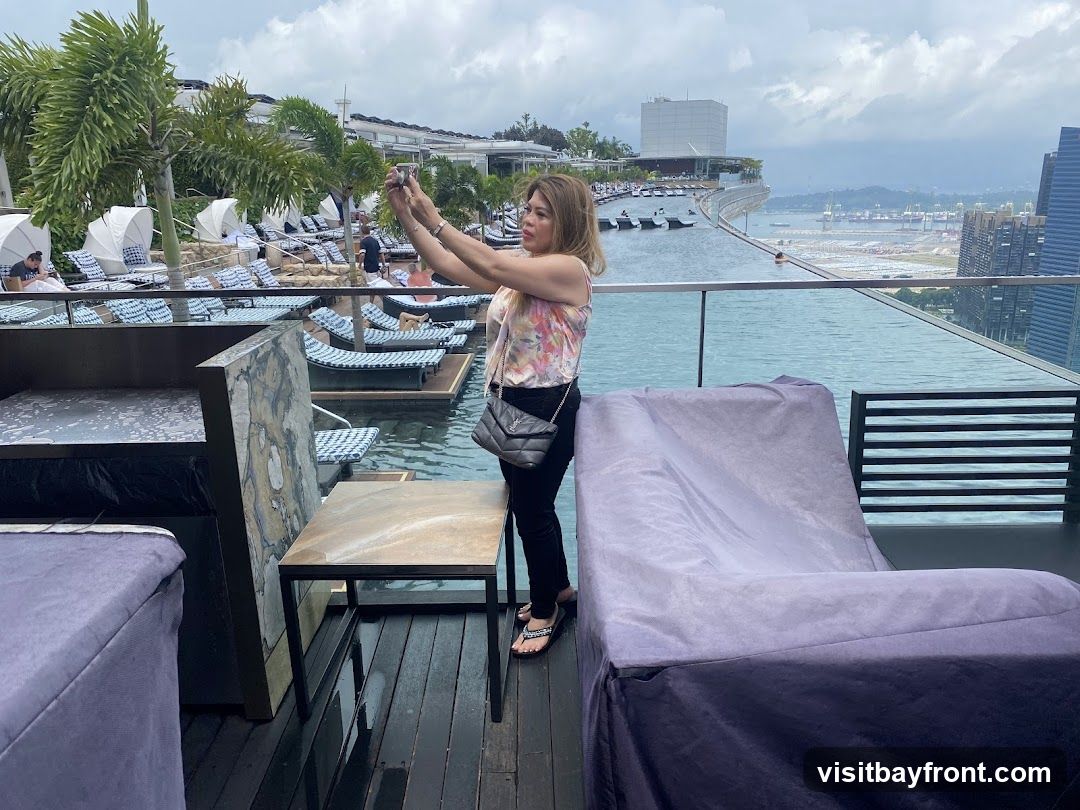 A woman stands on a wooden deck next to an infinity pool, holding up her phone to take a selfie. She is wearing a floral print top and black pants. In the background, there are rows of lounge chairs with white umbrellas, palm trees, and a cityscape with tall buildings. The pool overlooks a body of water and a distant port.