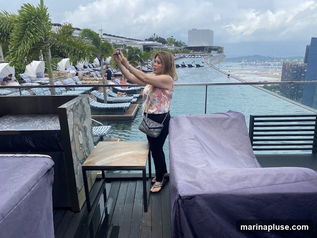 A woman stands on a wooden deck next to an infinity pool, holding up her phone to take a selfie. She is wearing a floral print top and black pants. In the background, there are rows of lounge chairs with white umbrellas, palm trees, and a cityscape with tall buildings. The pool overlooks a body of water and a distant port.