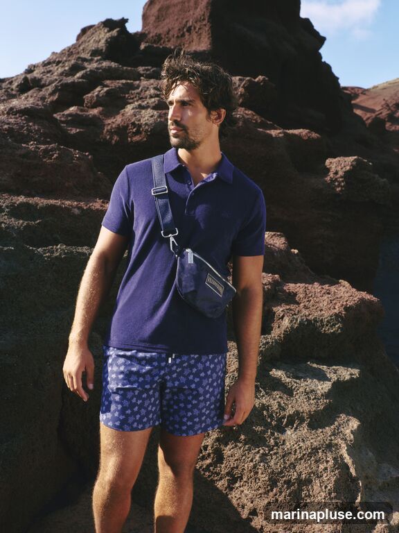 A man with dark hair and a beard stands on a rocky, reddish-brown terrain under a clear blue sky. He is wearing a dark blue polo shirt and patterned blue swim shorts. A small navy blue pouch is slung across his chest. The pouch has a rectangular label with text on it.