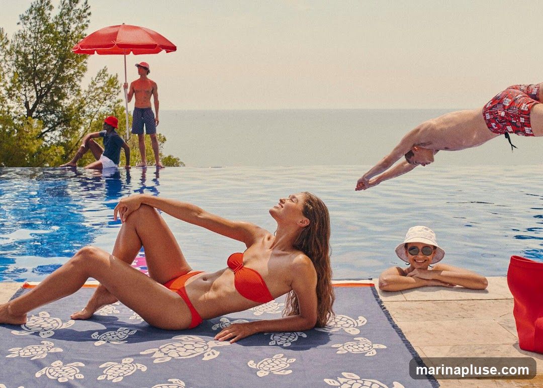 A group of people are enjoying a sunny day by an infinity pool. In the foreground, a woman in an orange bikini lies on a towel with a turtle pattern. To her right, a woman in a white bucket hat and sunglasses rests her arms on the pool edge. In the background, a man dives into the pool, while two other men stand near a red umbrella. The ocean is visible in the distance.
