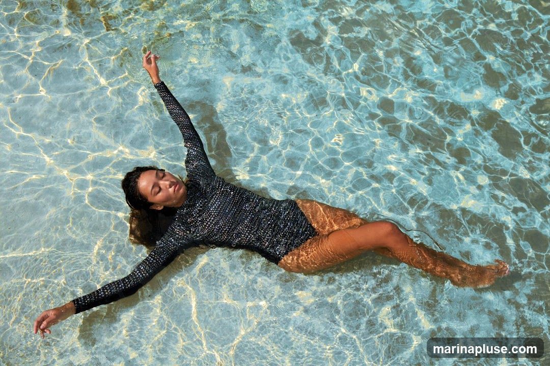 A woman with her eyes closed is floating on her back in a pool of clear blue water. She is wearing a long-sleeved black swimsuit with a textured pattern. Sunlight refracts through the water, creating shimmering patterns on her body and the pool floor.