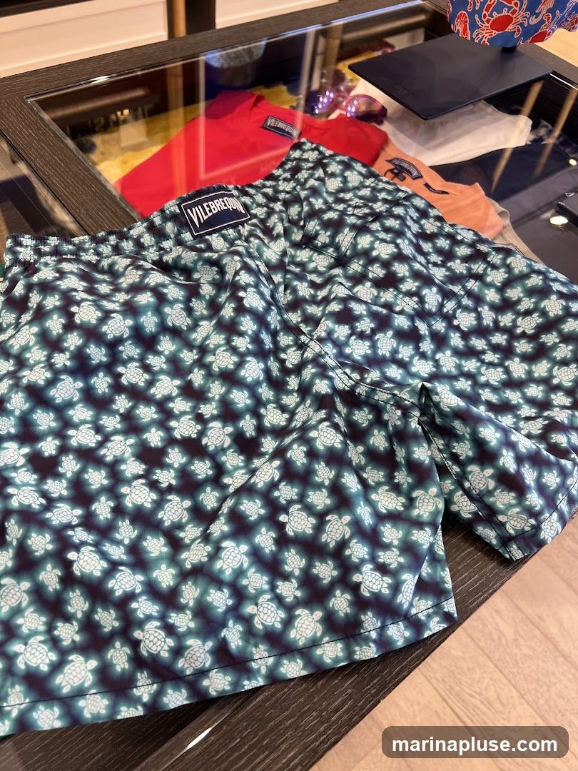 A pair of Vilebrequin swim trunks with a dark blue background and a pattern of light blue turtles. The trunks are laid out on a glass display table, with other clothing items visible in the background, including a red t-shirt and an orange t-shirt. A Vilebrequin logo tag is visible on the waistband of the swim trunks.