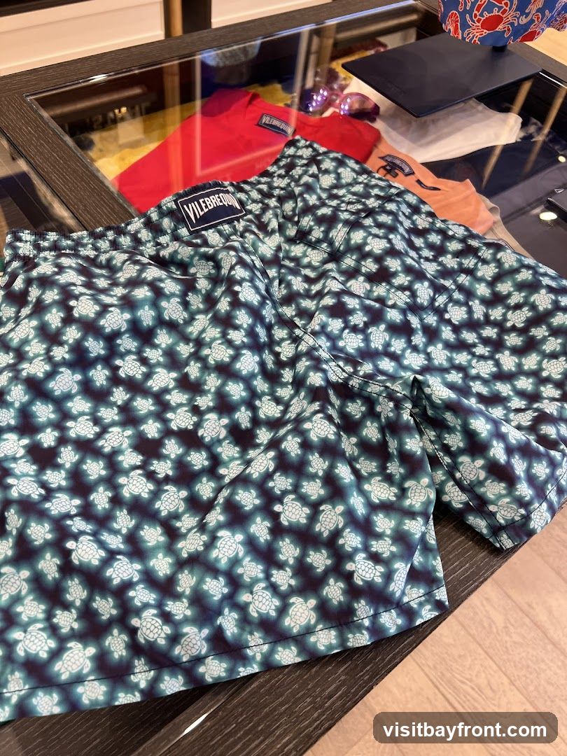 A pair of Vilebrequin swim trunks with a dark blue background and a pattern of light blue turtles. The trunks are laid out on a glass display table, with other clothing items visible in the background, including a red t-shirt and an orange t-shirt. A Vilebrequin logo tag is visible on the waistband of the swim trunks.