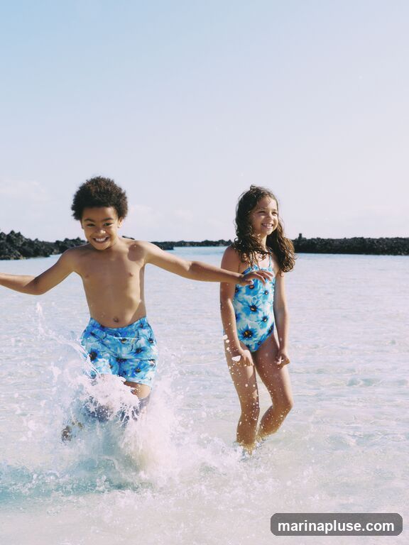 Two children, a boy and a girl, are running through shallow ocean water. The boy is wearing blue swim trunks with a floral pattern, and the girl is wearing a blue swimsuit with a floral pattern. They are both smiling and appear to be enjoying themselves. The water is clear and there are splashes around their legs. In the background, there is a clear blue sky and a dark, rocky shoreline.