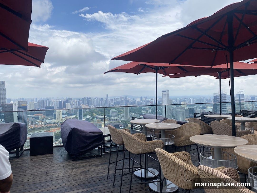 An outdoor seating area on a rooftop, featuring numerous tables and chairs with a panoramic view of a sprawling cityscape under a cloudy sky. Red umbrellas provide shade over some of the tables. The seating includes wicker chairs and bar stools. Some furniture is covered with dark tarps.