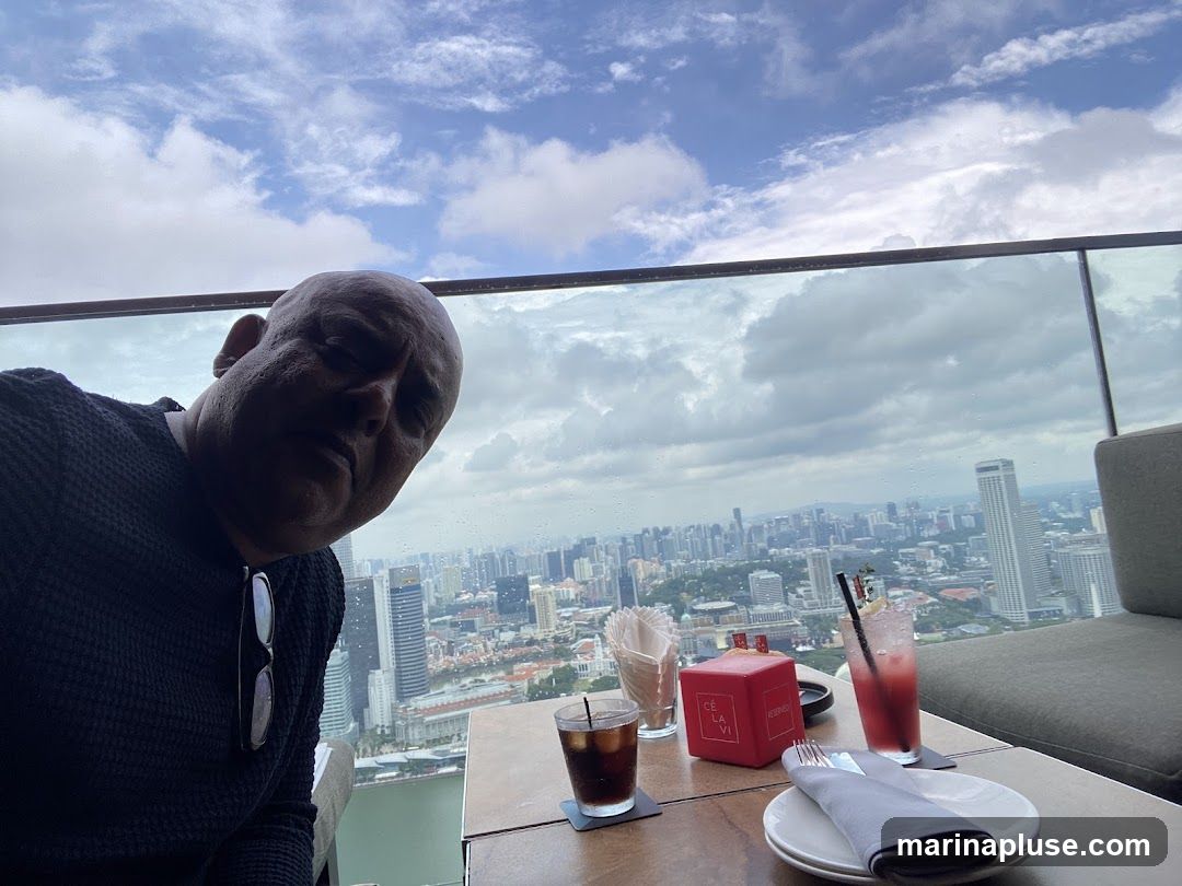 A man is taking a selfie at a rooftop bar, with a panoramic view of a city skyline in the background. He is wearing a dark sweater and has sunglasses clipped to his shirt. On the table in front of him are two drinks, a red cube with the text "CE LA VI RESERVED" on it, and a plate with cutlery. The sky is cloudy.