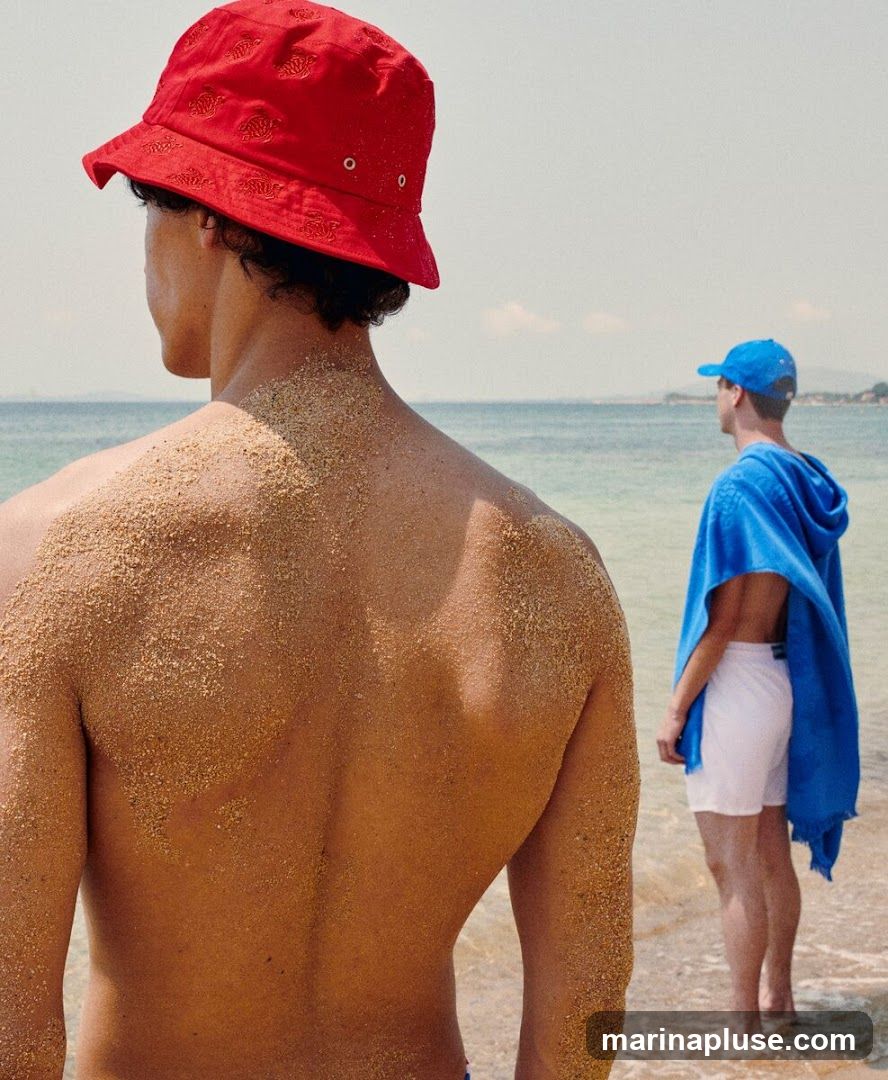 A man in the foreground, facing away from the camera, is covered in sand. He is wearing a red bucket hat. In the background, another man, also facing away, is wearing white shorts and a blue hooded towel draped over his shoulders. He is also wearing a blue baseball cap. They are standing on a sandy beach with the ocean and a distant coastline visible.