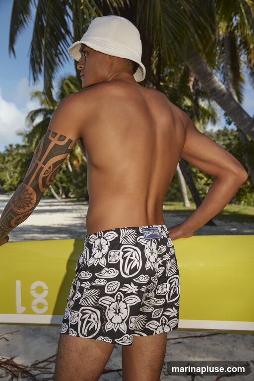 A man wearing a white bucket hat and black and white patterned swim trunks stands on a sandy beach, with palm trees and a bright blue sky in the background. He is holding a yellow object, possibly a surfboard or paddleboard, and has a tribal tattoo on his left arm.