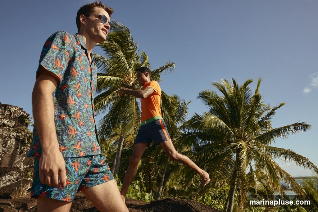 Two men are posing outdoors with palm trees and a clear blue sky in the background. The man in the foreground is wearing a short-sleeved button-down shirt and shorts with a tropical floral print in shades of teal, orange, and green. He is also wearing sunglasses. The man in the background is wearing an orange t-shirt and navy blue swim trunks with an orange stripe. He is in motion, with one leg extended. The setting appears to be a tropical or subtropical location with lush vegetation and rocky terrain.