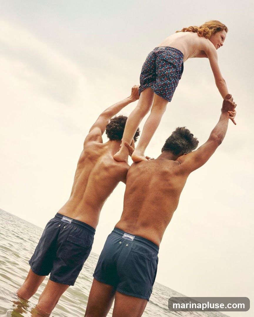 Three men are in the ocean, with two men supporting a third man who is standing on their shoulders. The men are wearing swim trunks. The sky is overcast.