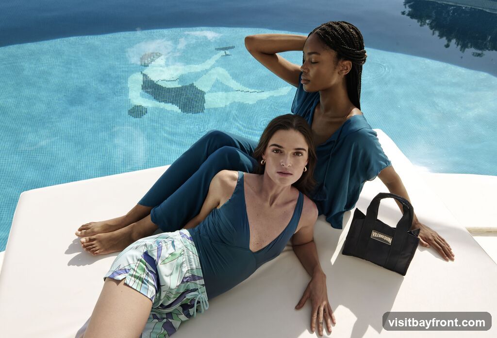 Two women are relaxing by a swimming pool. The woman in the foreground is wearing a teal swimsuit and patterned shorts. The woman behind her is wearing a teal top and pants. A black tote bag with the Vilebrequin logo is placed next to them. The pool has a mosaic design at the bottom.
