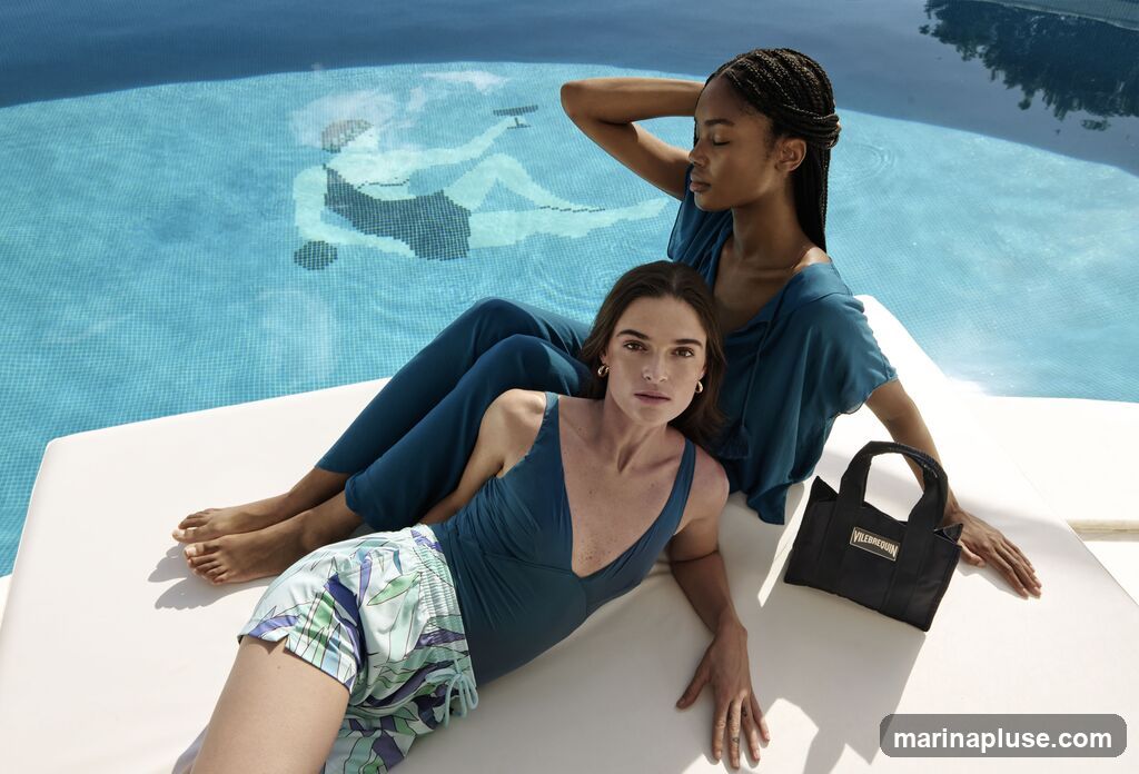 Two women are relaxing by a swimming pool. The woman in the foreground is wearing a teal swimsuit and patterned shorts. The woman behind her is wearing a teal top and pants. A black tote bag with the Vilebrequin logo is placed next to them. The pool has a mosaic design at the bottom.