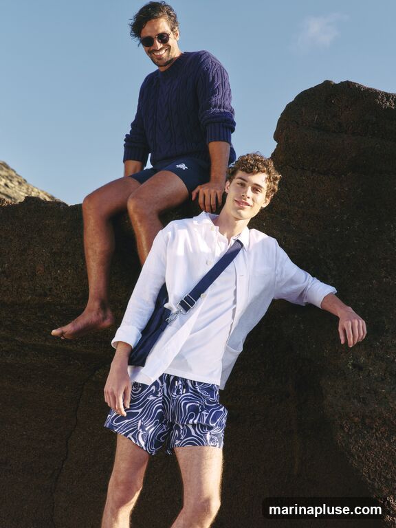 Two men are posing on large rocks outdoors. The man in the foreground is wearing a white open-collared shirt over a white t-shirt, and blue and white patterned swim shorts. He has a blue messenger bag slung across his body. The man behind him is wearing a navy blue cable-knit sweater and navy blue swim shorts with a small white logo. He is also wearing sunglasses and is barefoot. The sky is clear and blue.