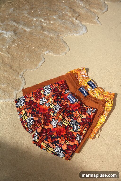 Three pairs of swim trunks are laid out on a sandy beach with gentle waves lapping at the shore. The trunks feature vibrant patterns, including floral designs and sea turtles. One pair has a dark blue base with red and orange flowers and white sea turtles. Another pair is a solid rust color. The third pair has a bright orange and yellow abstract pattern. Each pair has a visible blue tag with white lettering that reads "VILEBREQUIN".