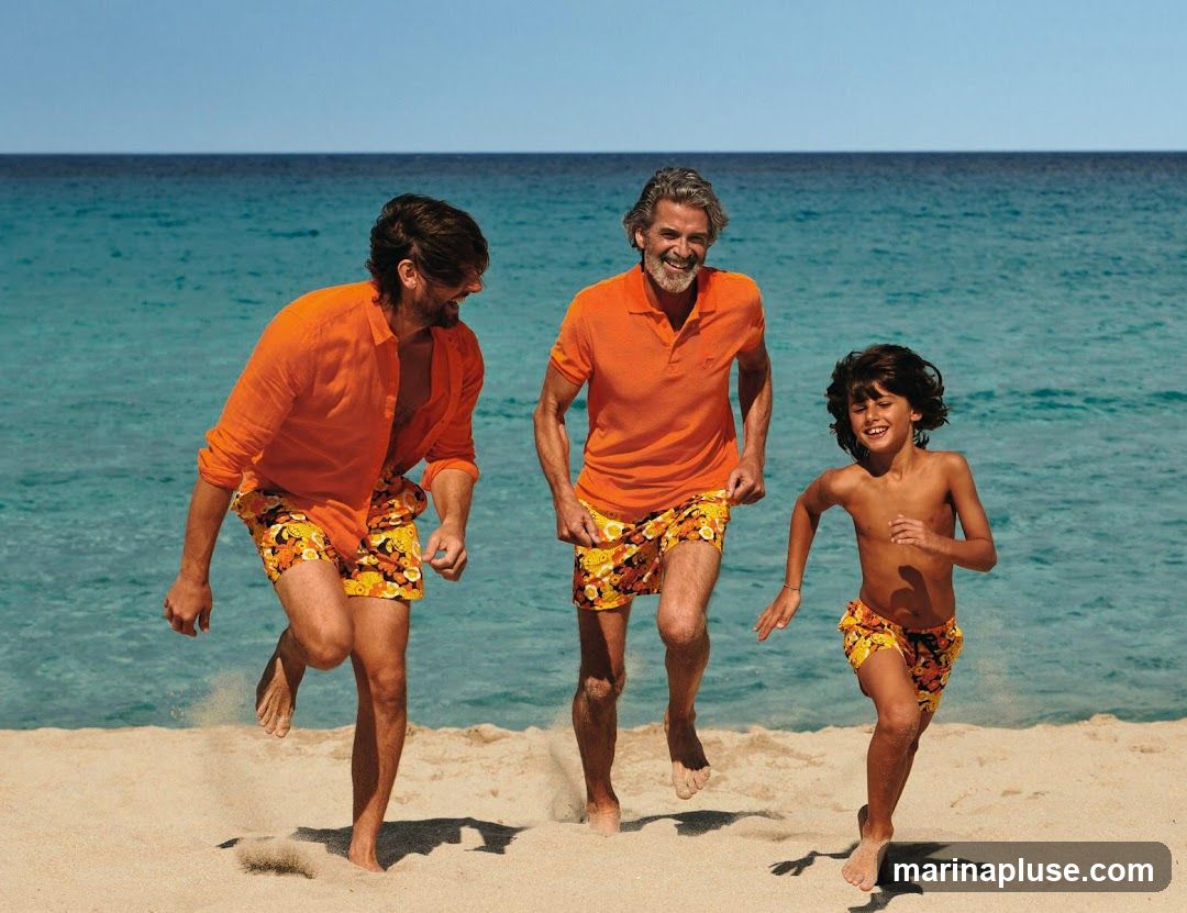 Three people, two men and a boy, are running on a sandy beach with the ocean in the background. They are all wearing orange shirts and patterned swim trunks. The man on the left is wearing a long-sleeved orange shirt and floral swim trunks. The man in the middle is wearing an orange polo shirt and floral swim trunks. The boy on the right is shirtless and wearing floral swim trunks. They are all smiling and appear to be enjoying themselves.