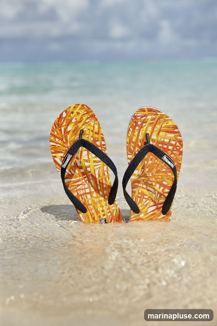 A pair of orange and yellow patterned flip-flops are partially submerged in shallow water on a sandy beach. The ocean and sky are visible in the background.