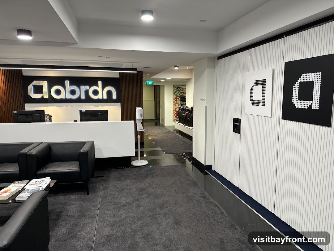 A modern office reception area with a prominent "abrdrn" logo illuminated behind a white reception desk. Black leather seating is arranged in the foreground, with magazines on a coffee table. To the right, the "abrdrn" logo is displayed on a textured wall. An exit sign is visible down a hallway in the background.