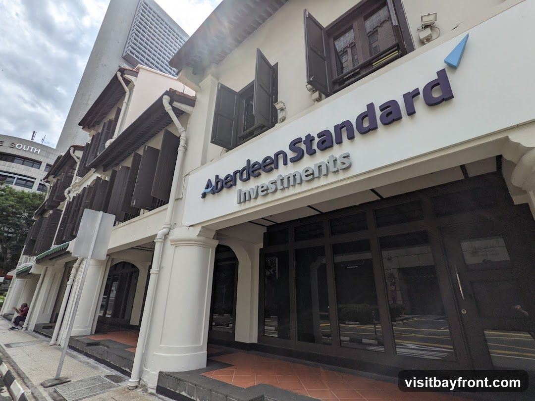 The exterior of a building with the sign "Aberdeen Standard Investments" prominently displayed. The building has traditional architecture with shutters on the windows, contrasting with a modern skyscraper in the background.