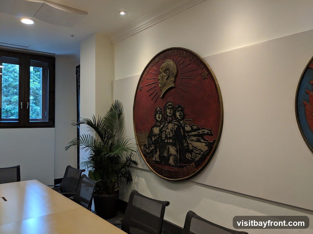 A conference room with a large circular medallion on the wall featuring a portrait of a man and a group of people. There is a large wooden table and chairs in the foreground, and a window with greenery visible outside.