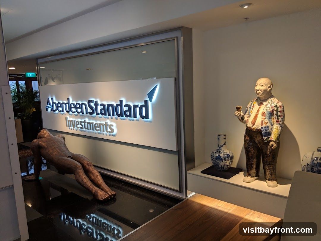 The image shows an interior view of an office entrance with the "Aberdeen Standard Investments" logo prominently displayed on a glass partition. To the left, a bronze sculpture of a reclining figure is visible. To the right, a statue of a man holding a drink stands next to decorative vases. An "EXIT" sign is also visible in the background.