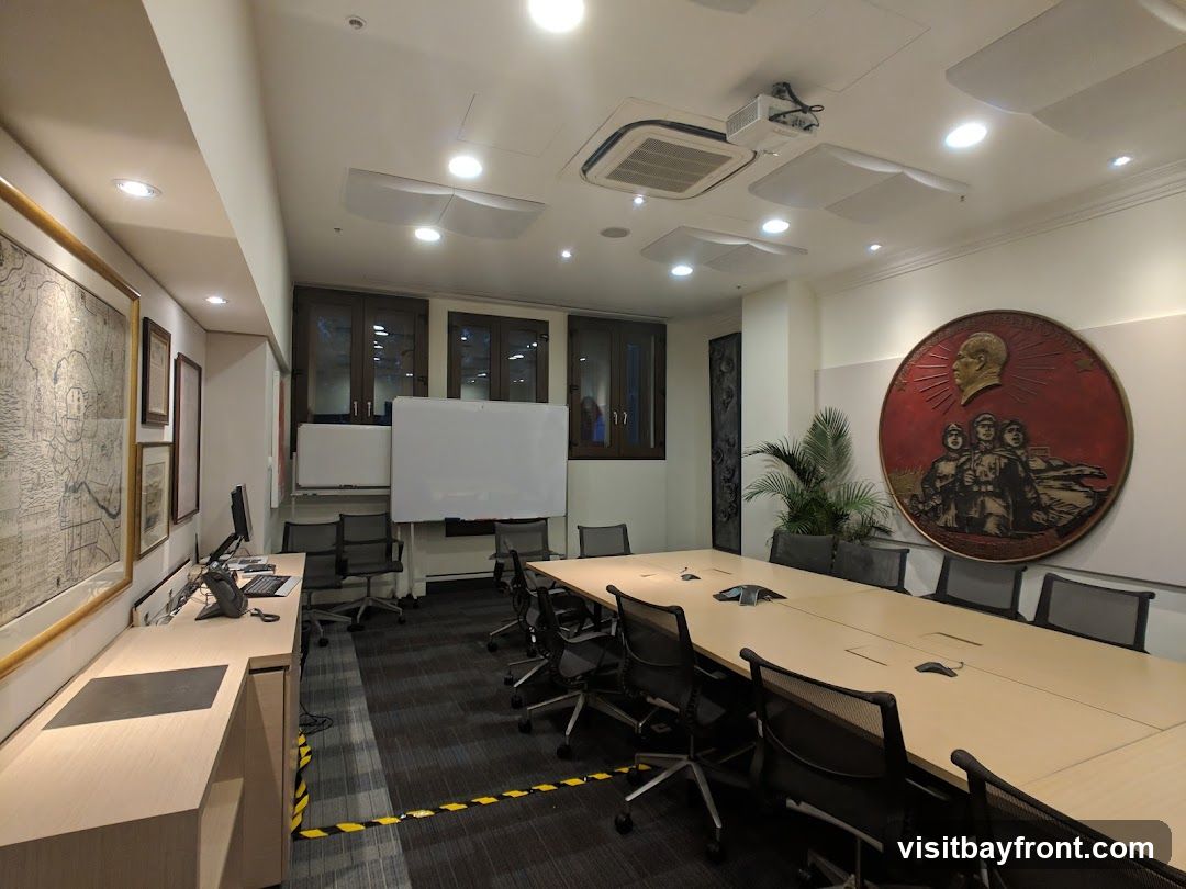 A well-lit conference room with a long table, chairs, whiteboards, and artwork on the wall. A projector is mounted on the ceiling. There is a desk area with a computer and phone.