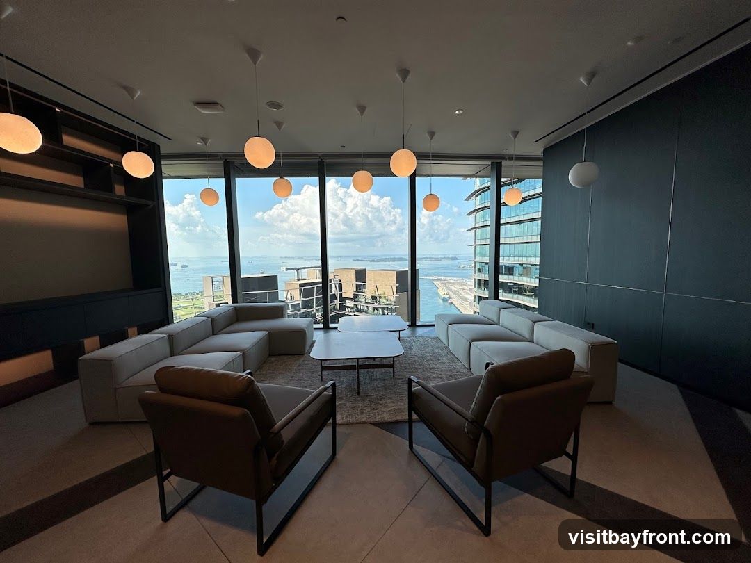 A contemporary lounge area with large windows offering a panoramic view of a city skyline and harbor. The room features modular sofas, armchairs, and a coffee table, all arranged for comfortable seating. Spherical pendant lights hang from the ceiling, illuminating the space. The interior design is sleek and minimalist, with dark walls and built-in shelving.