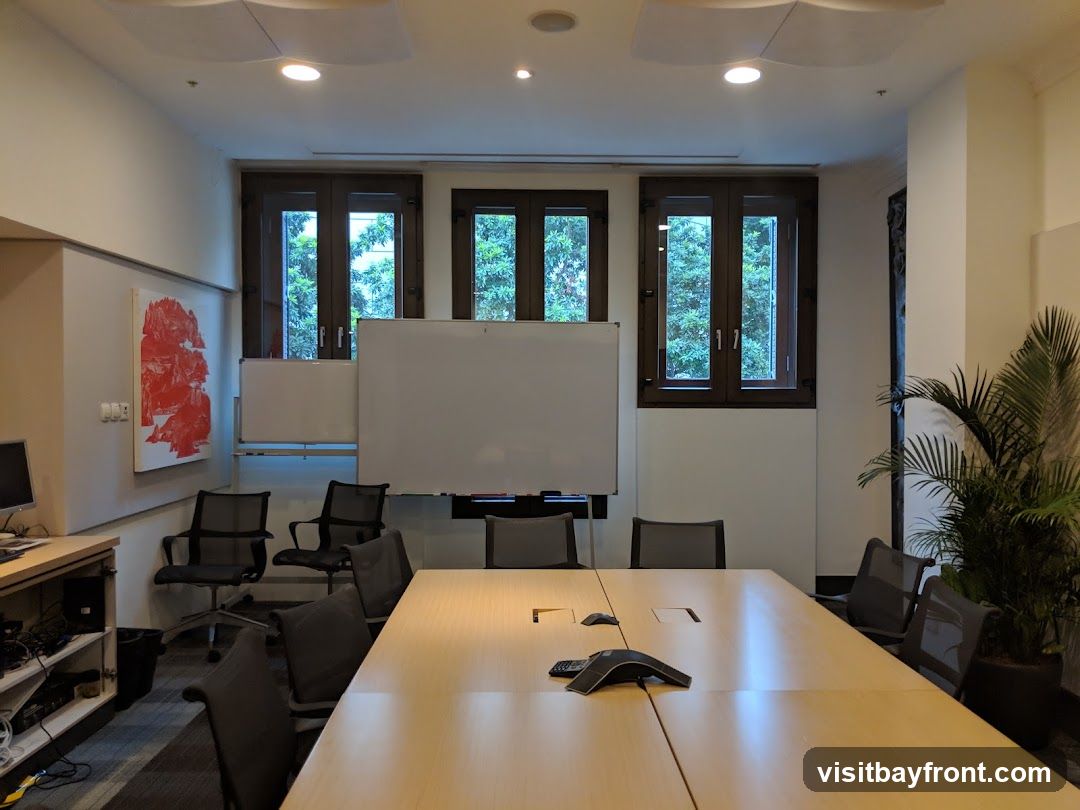 A modern conference room with a long wooden table, chairs, whiteboards, and windows overlooking trees. There is also a plant in a pot and a desk with computer equipment.