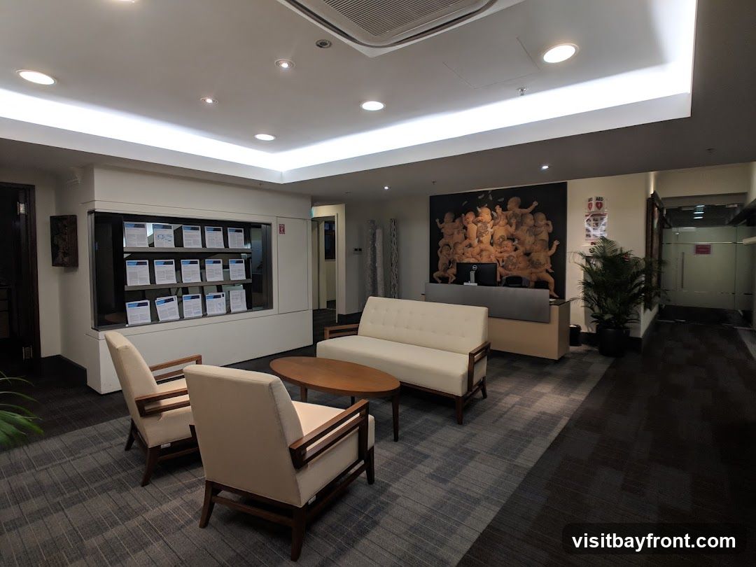 A modern office waiting area with comfortable seating, a reception desk, and artwork on the wall. There are two cream-colored armchairs and a matching sofa arranged around a wooden coffee table. Behind the sofa is a reception desk with a computer monitor and a large mural of cherubs. To the right of the desk, a potted plant and a sign are visible. A display case with documents is on the left wall.