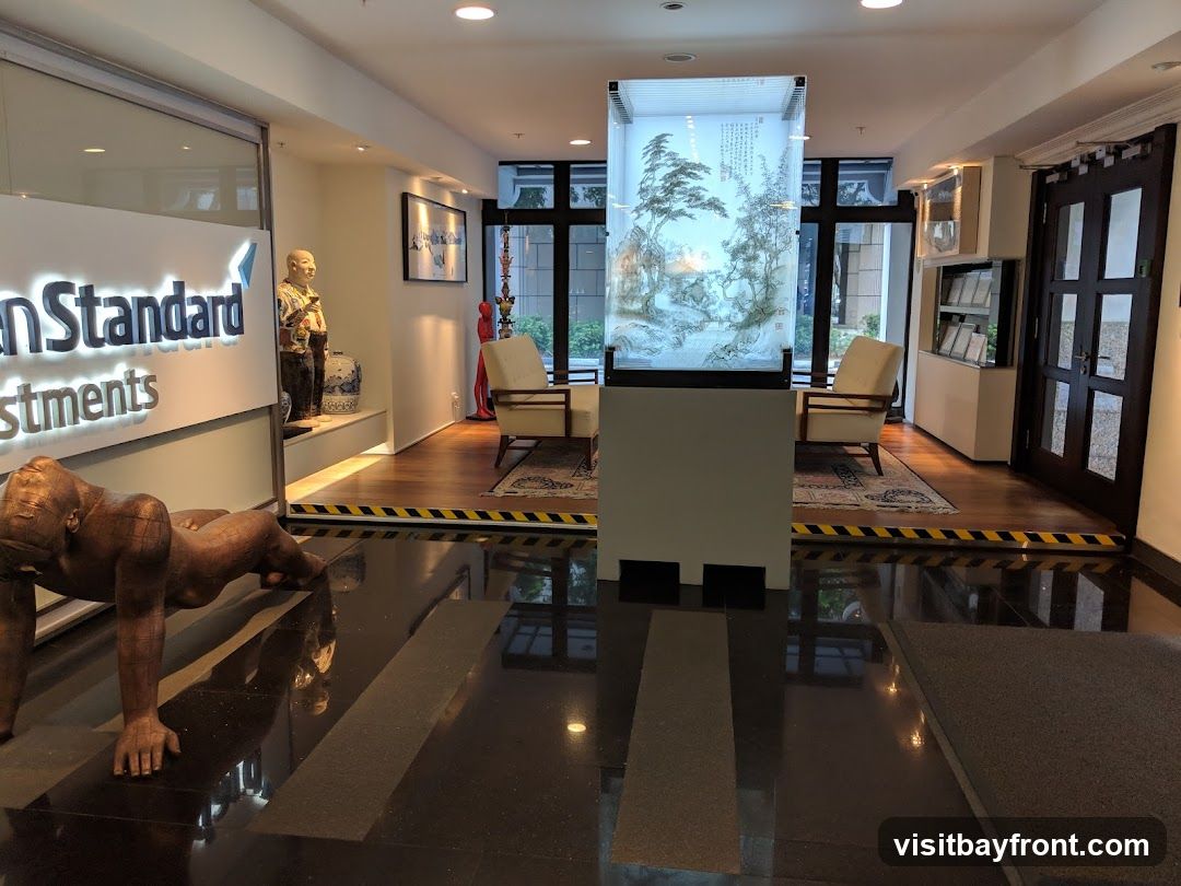 The image shows the interior of a modern office lobby. A large illuminated sign with the text "an Standard Investments" is visible on the left. In the center, there is a tall glass display case with an Asian-inspired artwork. Two armchairs are placed on a rug in front of the display. The floor is polished black granite, reflecting the lights and decor. A sculpture of a person is positioned in the foreground. The overall ambiance is sophisticated and professional.