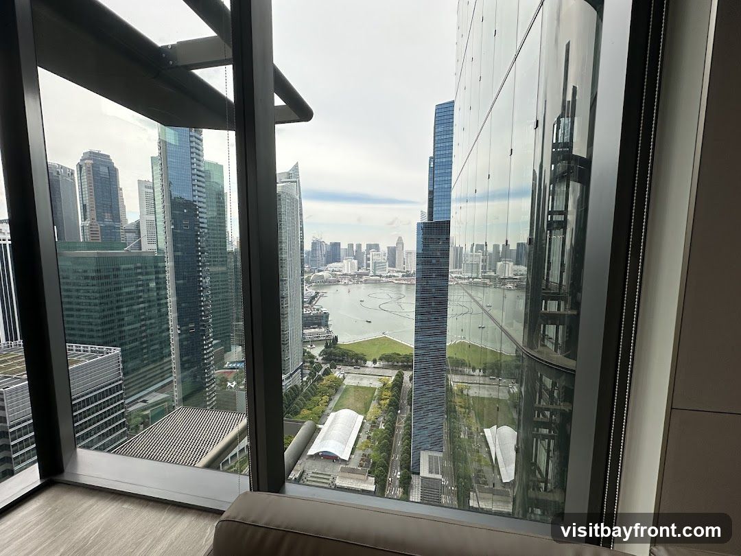 A panoramic view of a modern cityscape with numerous skyscrapers and a body of water, seen through a large window. The foreground shows the interior of a room with a couch and wooden flooring. The window frames the view, with reflections visible on the glass.