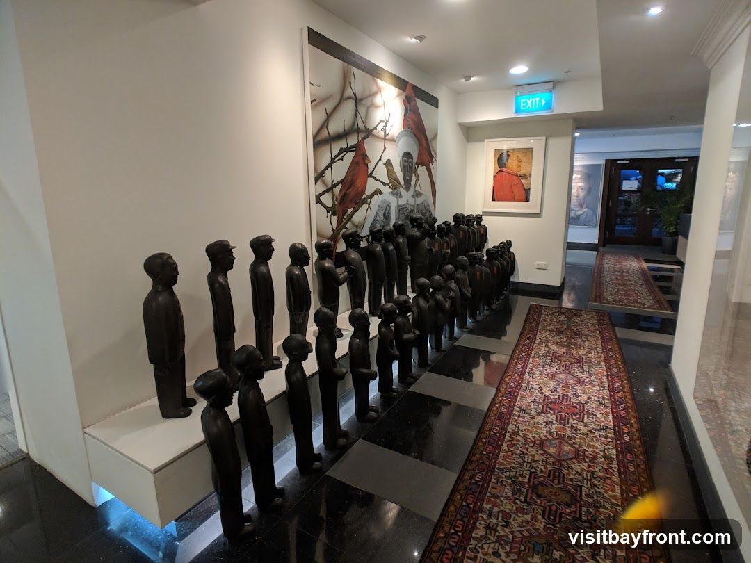 A hallway features a large art installation with numerous dark-colored human figurines arranged in rows. A large painting depicting birds and a figure hangs on the wall behind the figurines. Another framed artwork is visible on the adjacent wall, along with an illuminated "EXIT" sign. Two oriental rugs are laid out on the floor, leading towards a set of double doors at the end of the hallway.