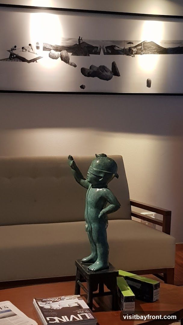 A bronze sculpture of a blindfolded child stands on a small stool in front of a sofa. Above the sofa, a long, black-framed artwork displays a surreal collage of landscapes, figures, and floating stones. On the coffee table in the foreground, magazines are stacked, and two green-topped boxes are placed.
