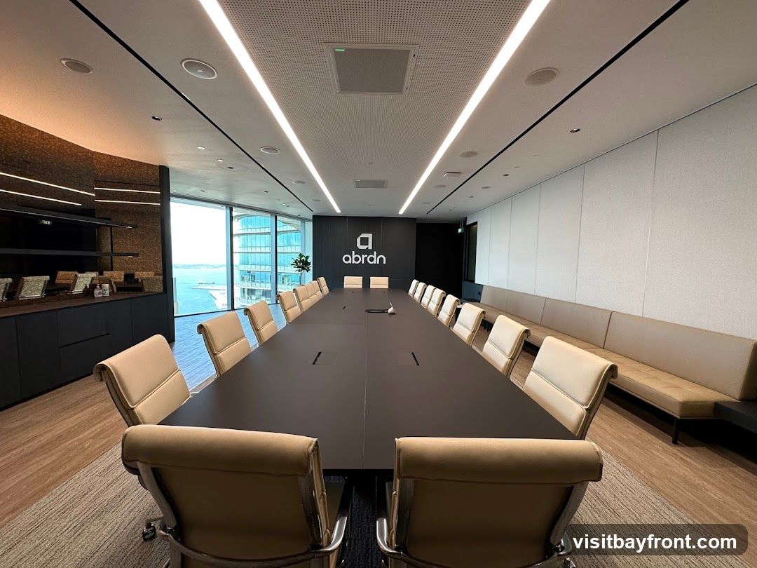 A spacious and modern conference room with a long, dark conference table surrounded by upholstered chairs. The room features large windows with a view of a city skyline and water. A branded logo is visible on the wall behind the table. The room is well-lit with linear ceiling lights and has a long bench seat along one wall.