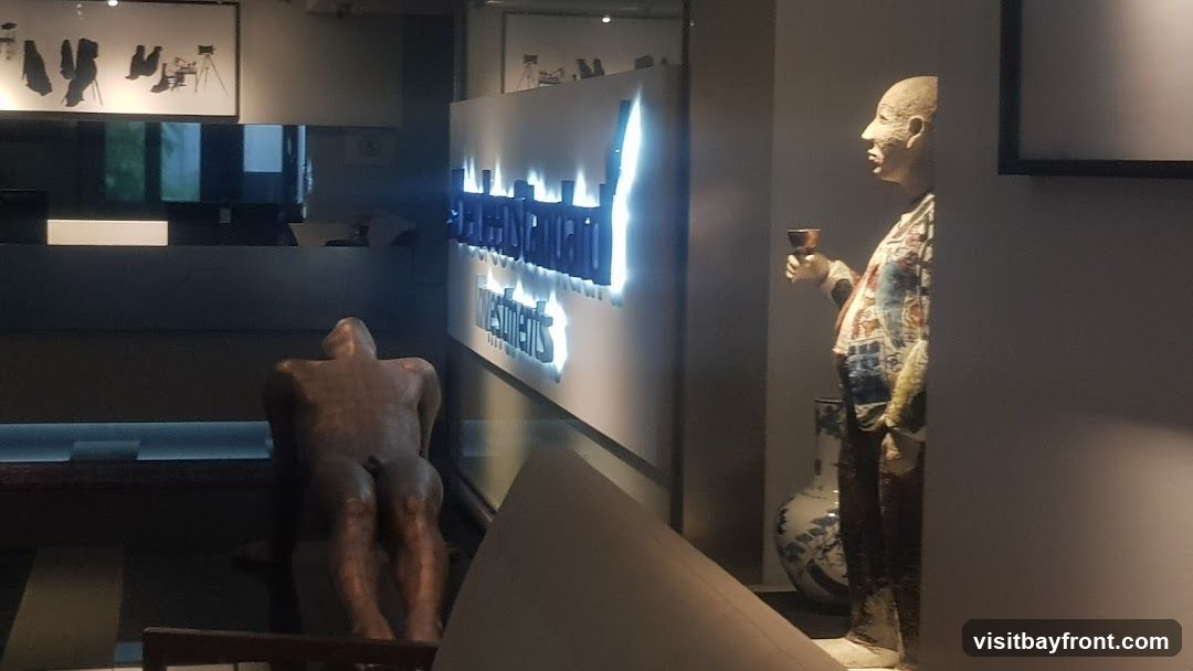 The image shows the interior of a building with a prominent illuminated sign that reads "WESTINGHAM INVESTMENTS". To the left, a bronze sculpture of a seated nude figure is visible. To the right, a colorful statue of a person holding a goblet stands next to a decorative vase. Artwork is displayed on the wall above the reception area.