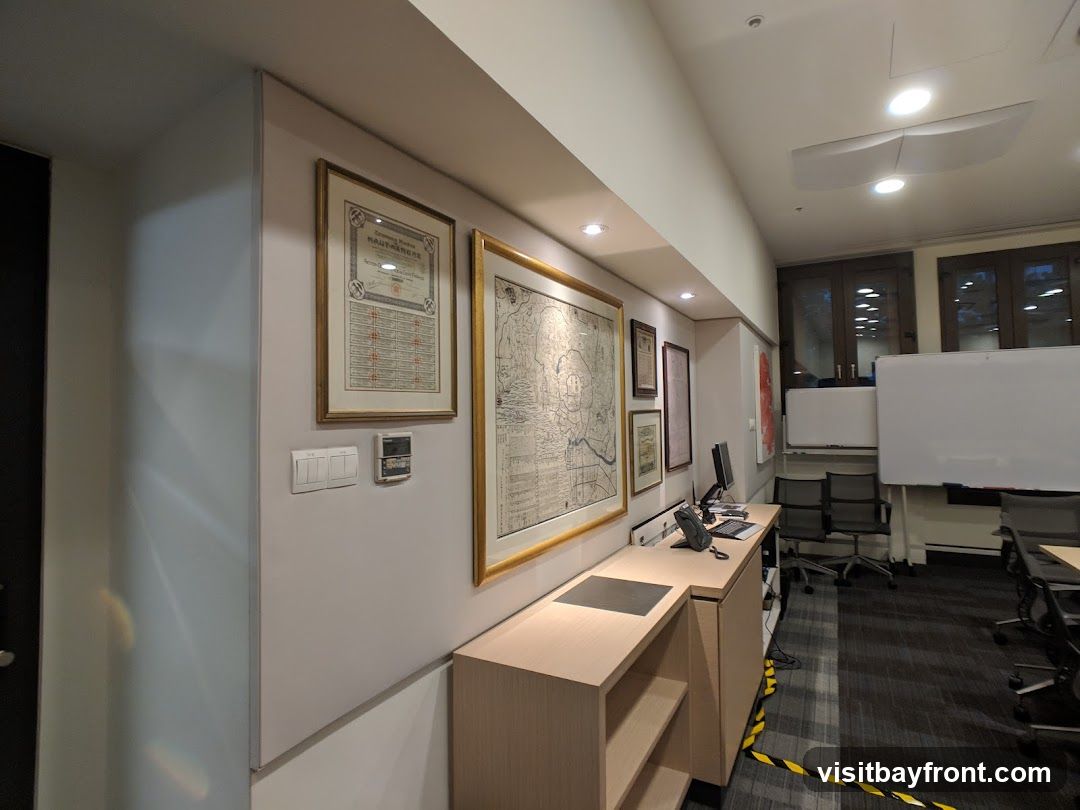 The image shows an office interior with several framed maps and documents on the wall. A desk with a computer monitor, telephone, and keyboard is visible. In the background, there are whiteboards, office chairs, and windows. The room appears to be a meeting or study space.