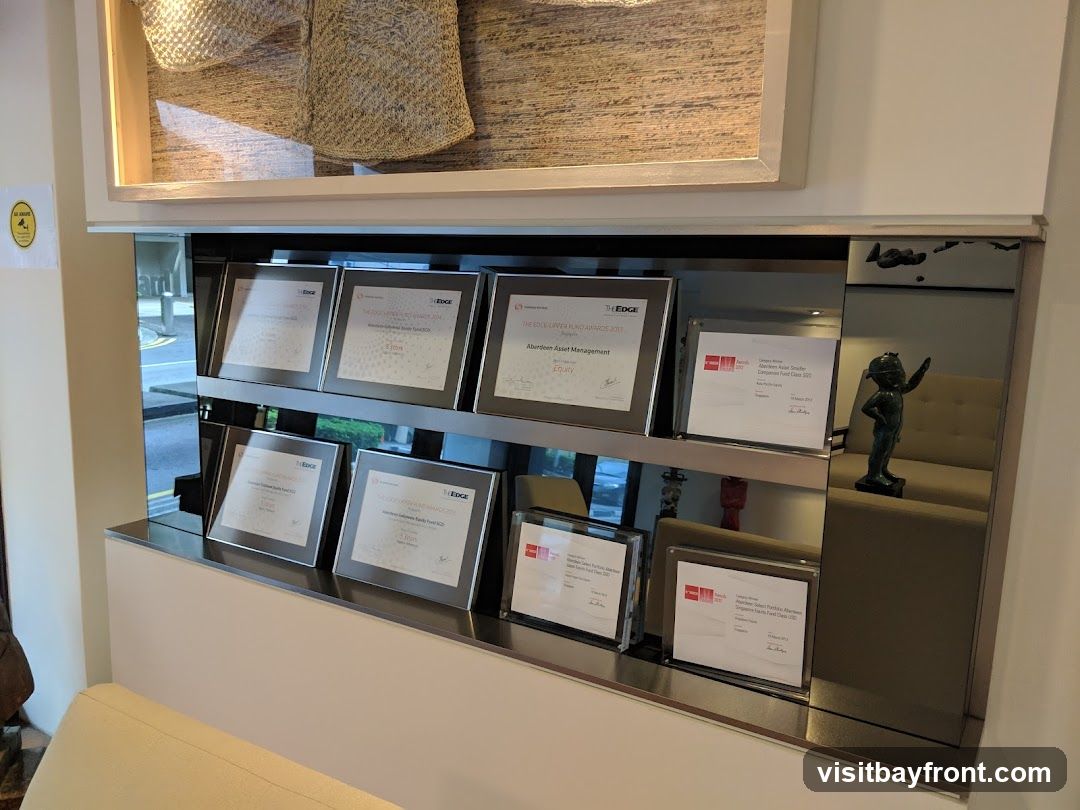 A display shelf featuring several framed awards and certificates, some with the logo "THE EDGE". A small bronze statue of a child is also visible. The awards appear to be related to asset management and funds. In the background, through a reflective surface, a street scene and a sofa can be glimpsed.
